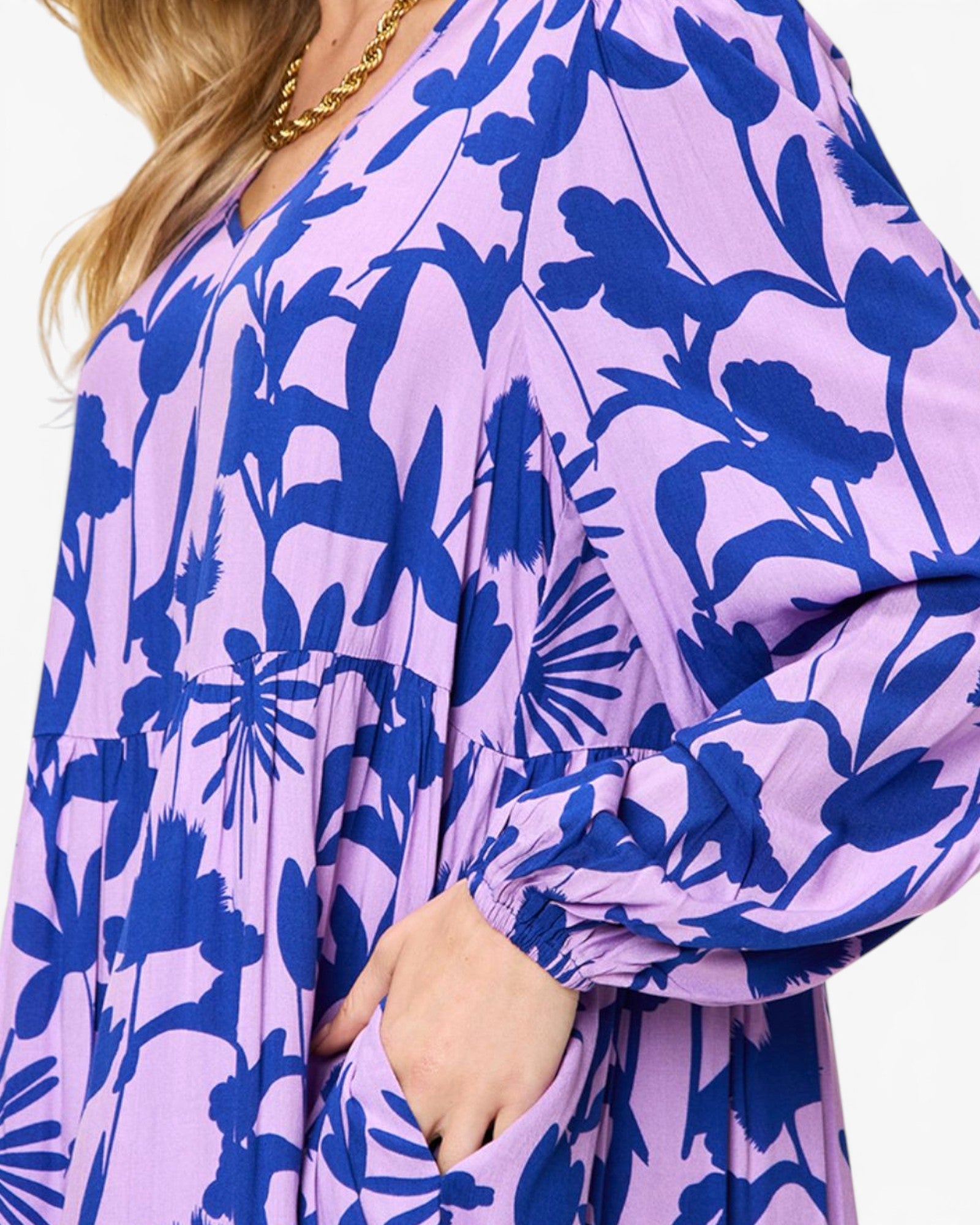 A close-up of a woman wearing a loose long-sleeve dress with a lavender purple and dark blue floral pattern, featuring a V-neckline and a gold chain necklace.
