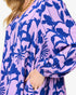 A close-up of a woman wearing a loose long-sleeve dress with a lavender purple and dark blue floral pattern, featuring a V-neckline and a gold chain necklace.