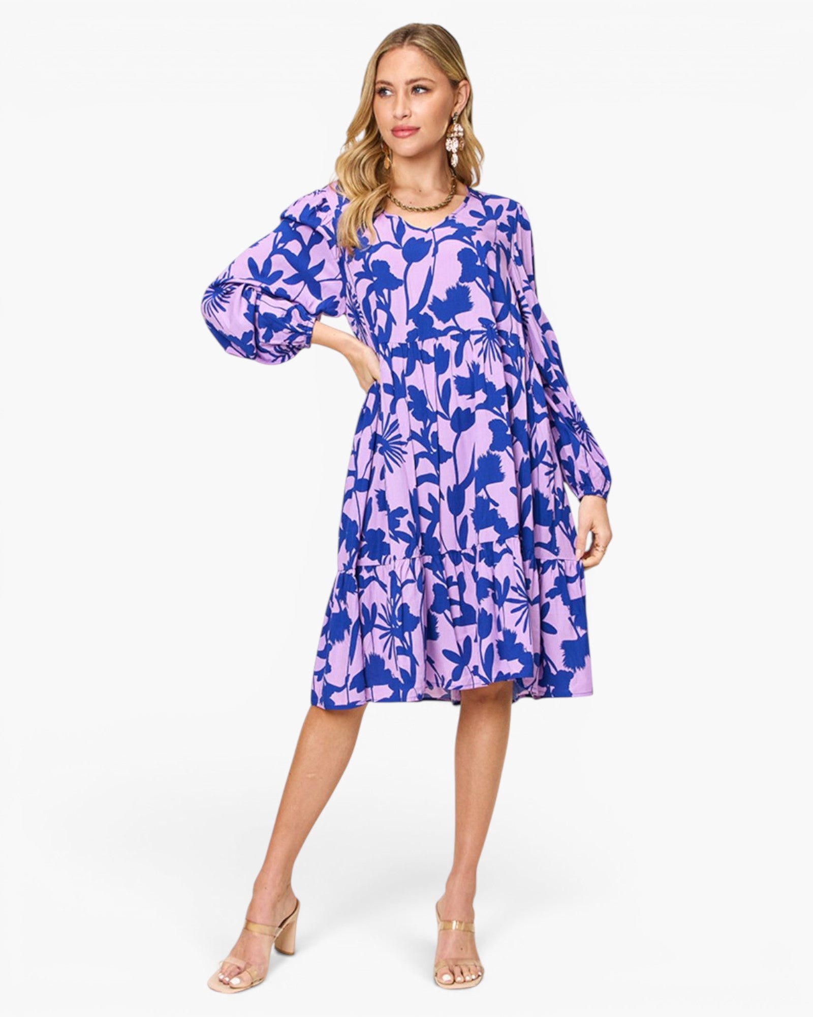 A woman modeling a knee-length tiered dress with a blue and lavender floral print, featuring voluminous long sleeves and a round neckline, set against a plain white studio background.