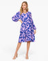 A woman modeling a knee-length tiered dress with a blue and lavender floral print, featuring voluminous long sleeves and a round neckline, set against a plain white studio background.