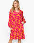 A woman modeling a long-sleeve tiered midi dress with a vibrant pink and orange floral pattern, set against a plain white studio background.