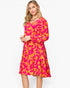 A woman modeling a long-sleeve knee-length dress with a bold abstract floral pattern in hot pink and orange against a clean white studio background.
