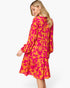 A woman modeling a long-sleeve midi dress with a vibrant pink and orange floral pattern, shown from a three-quarters back view against a plain white background.