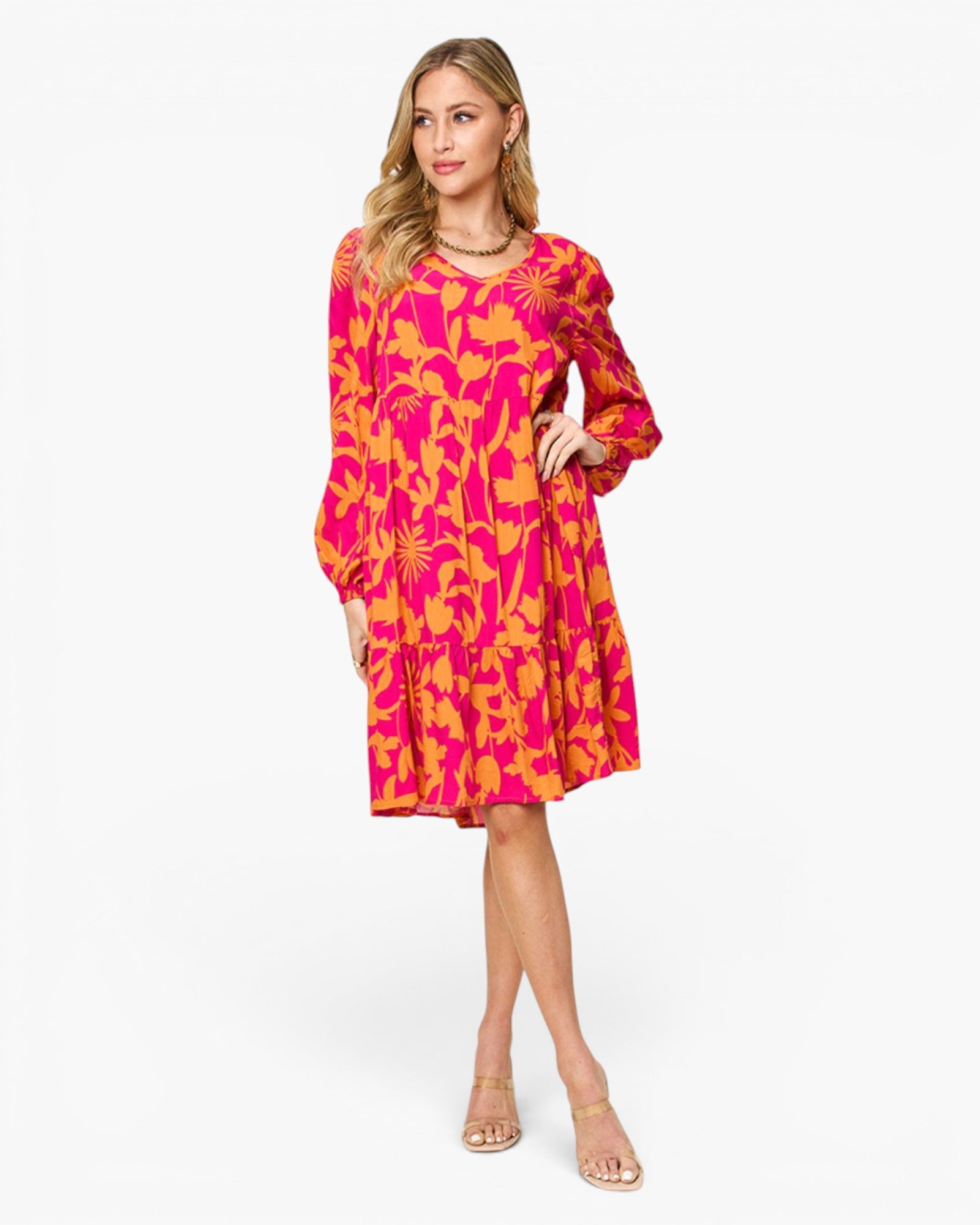 A woman modeling a bright pink and orange tropical print dress with long sleeves and a tiered skirt, paired with clear high-heel sandals, against a plain white studio background.