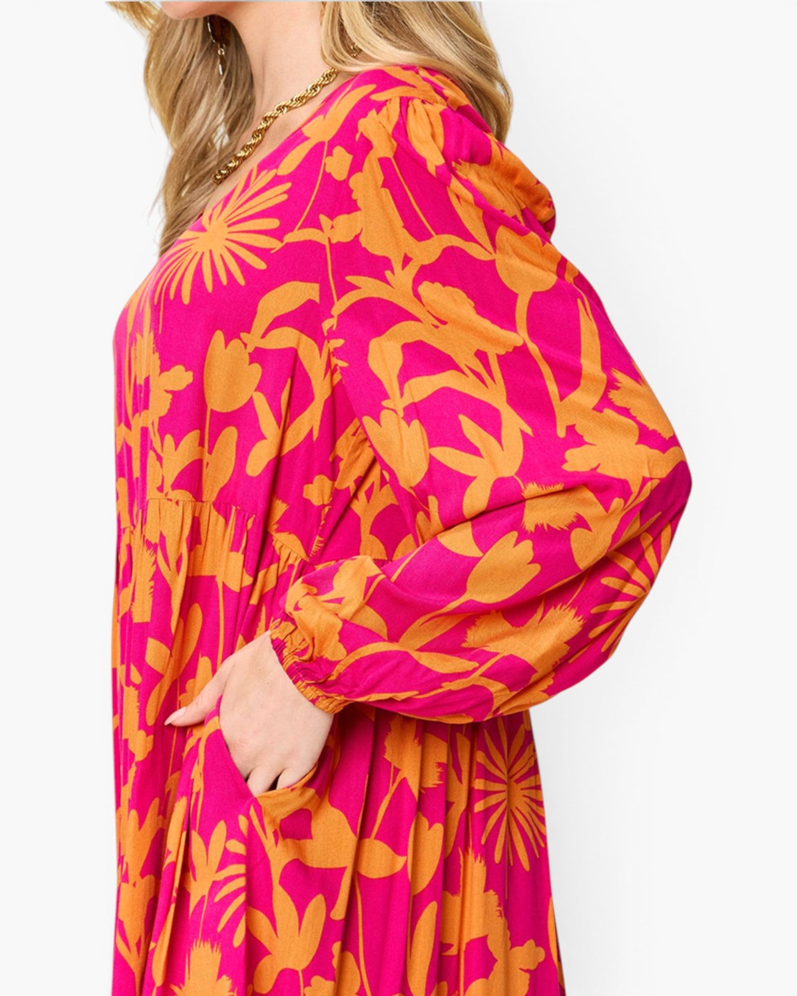 A close-up side view of a woman wearing a bright pink and orange floral print long-sleeve dress against a plain white background.
