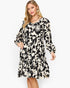 A woman modeling a long-sleeve tiered dress with a black and cream floral print against a plain white studio background.