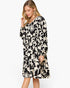 A woman modeling a long-sleeve V-neck dress with a black and cream floral and leaf pattern, set against a plain light gray background.