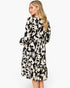 A woman modeling a black and cream floral print midi dress from the back, featuring long sleeves and a ruffled hem, against a plain light gray background.