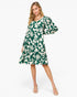 A woman modeling a knee-length green and white tropical leaf print dress with balloon sleeves, paired with clear high-heeled sandals, against a plain white studio background.
