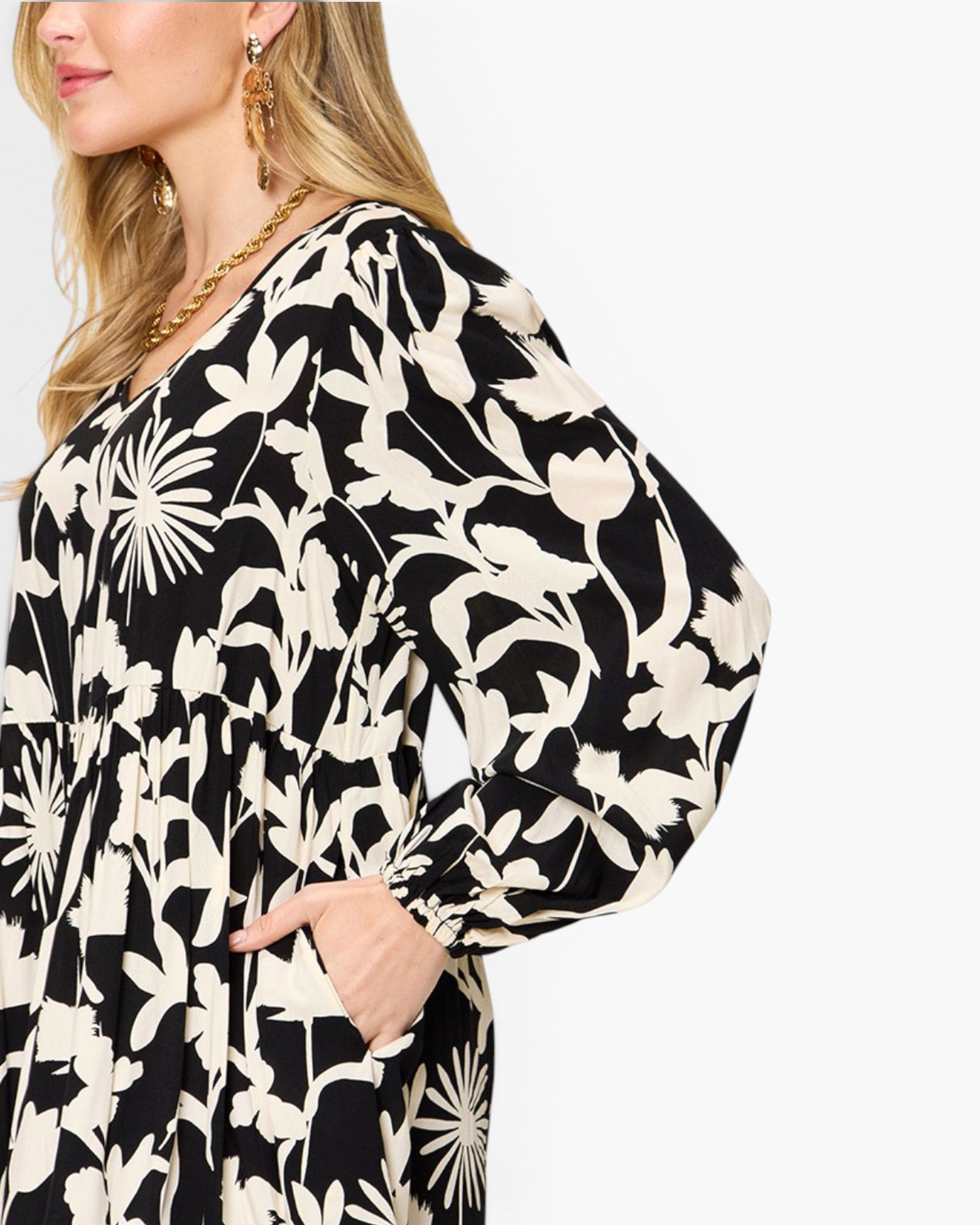 A close-up of a woman wearing a black and cream floral print dress with voluminous balloon sleeves and side pockets, set against a plain light gray background.