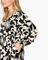A close-up of a woman wearing a black and cream floral print dress with voluminous balloon sleeves and side pockets, set against a plain light gray background.