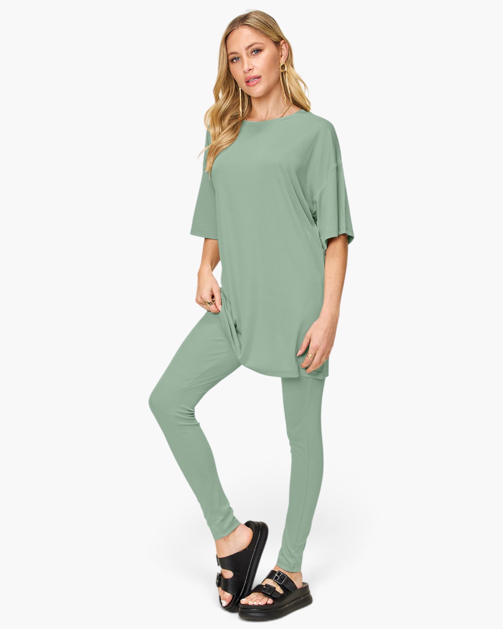 A woman modeling a matching sage green lounge set consisting of a short-sleeve tunic top and high-waisted leggings, paired with black slide sandals, against a plain white studio background.