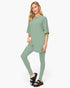 A woman modeling a matching sage green lounge set consisting of a short-sleeve tunic top and high-waisted leggings, paired with black slide sandals, against a plain white studio background.