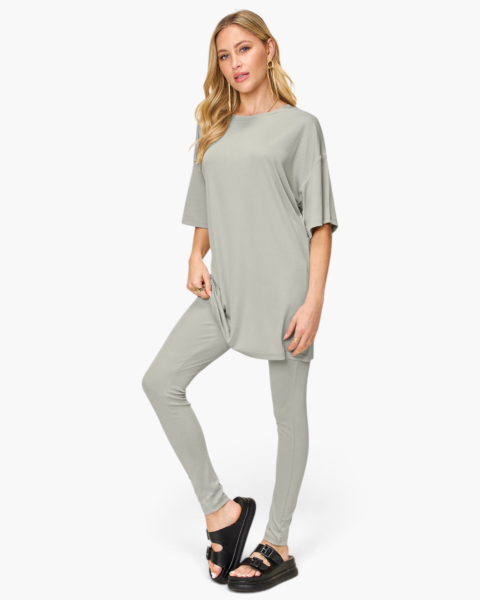 A woman modeling a light sage green matching lounge set consisting of a short-sleeve tunic top and high-waisted leggings, paired with black slide sandals, against a plain white studio background.