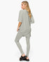 A woman modeling a light sage green two-piece lounge set consisting of an oversized short-sleeve top and matching leggings, shown from a side-back angle against a plain white studio background.