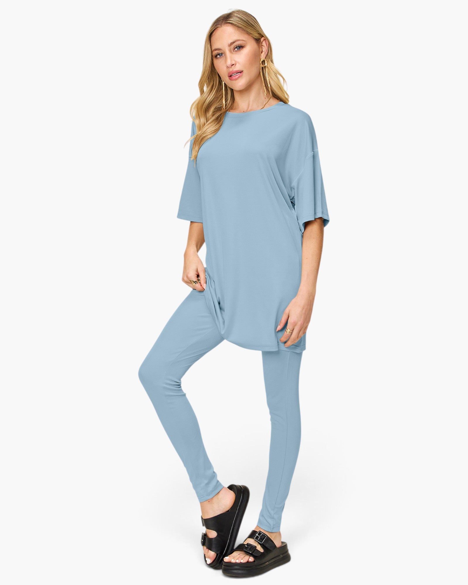 A woman modeling a light blue matching lounge set consisting of a short-sleeve tunic top and high-waisted leggings, paired with black slide sandals, against a plain white studio background.