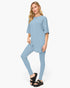 A woman modeling a light blue matching lounge set consisting of a short-sleeve tunic top and high-waisted leggings, paired with black slide sandals, against a plain white studio background.