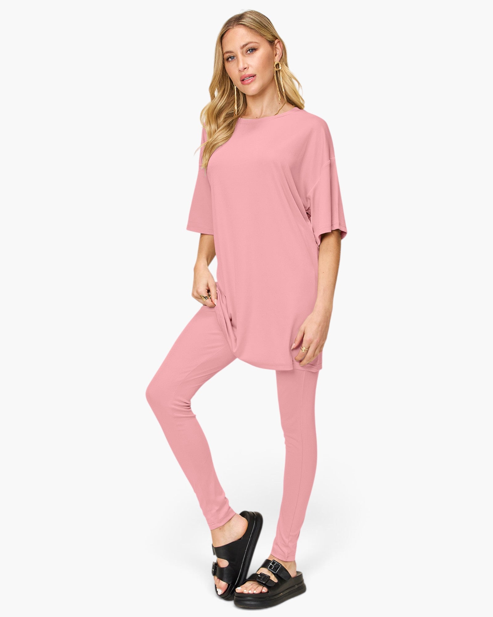 A woman modeling a matching pink lounge set consisting of a short-sleeve tunic top and high-waisted leggings, paired with black slide sandals, against a plain white studio background.