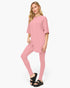 A woman modeling a matching pink lounge set consisting of a short-sleeve tunic top and high-waisted leggings, paired with black slide sandals, against a plain white studio background.