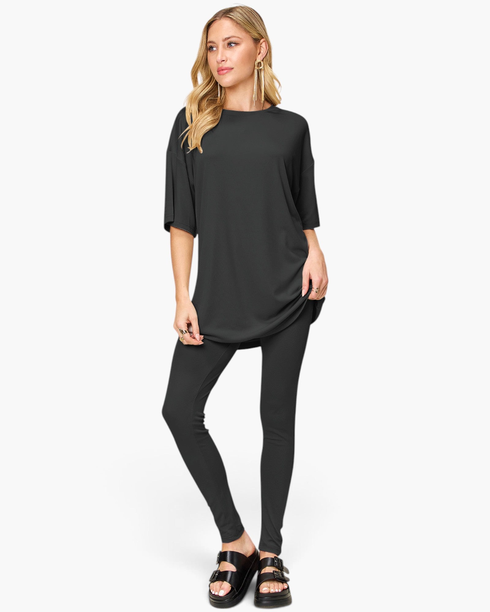 A woman modeling a black oversized short-sleeve tunic top paired with black leggings and black sandals, standing against a plain light gray studio background.
