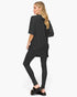 A woman modeling a black oversized short-sleeve tunic top with matching black leggings and black slide sandals, shown from a side-back angle against a plain white studio background.