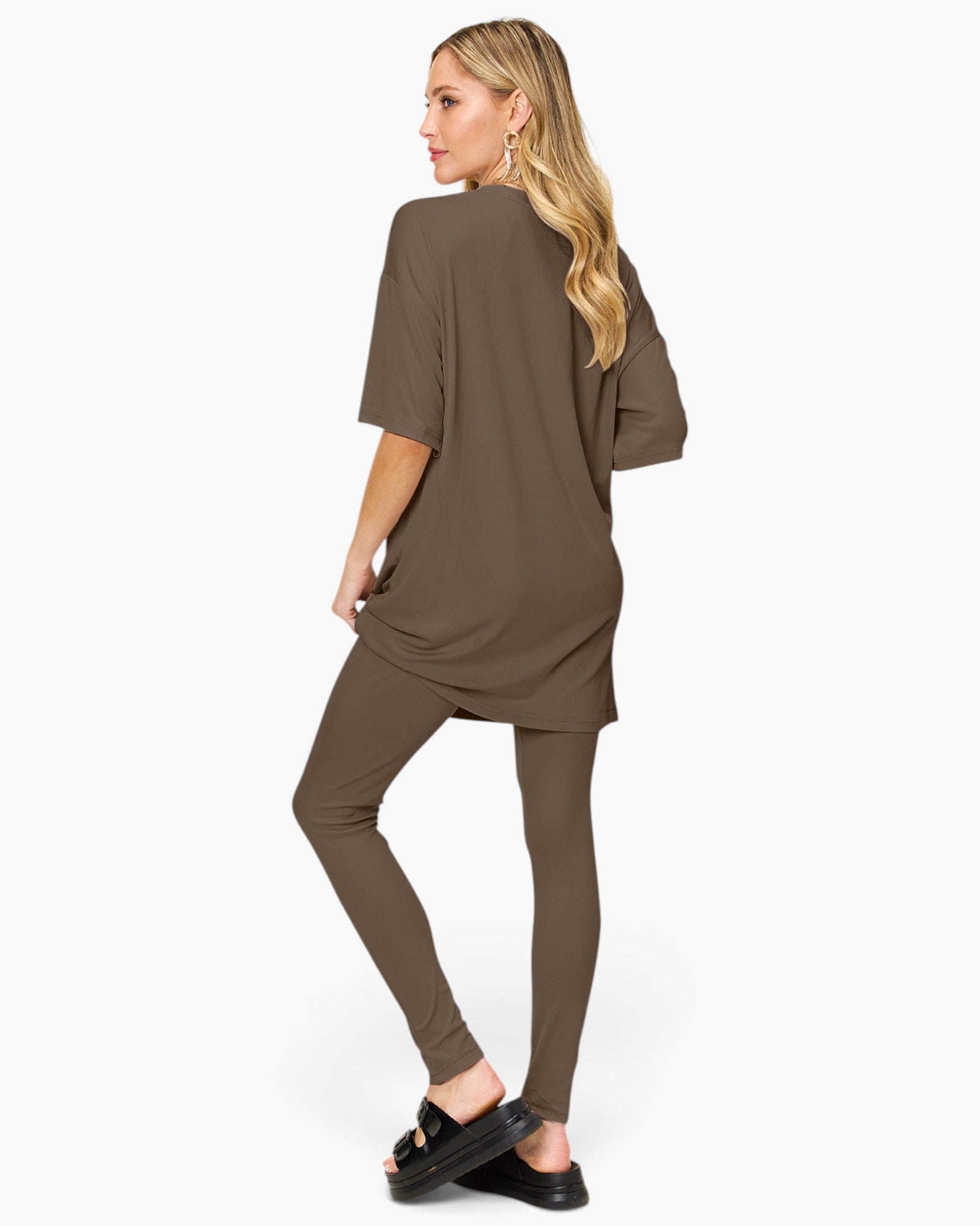 A woman modeling a matching two-piece lounge set consisting of an oversized short-sleeve tunic top and high-waisted leggings in a solid taupe-brown color, shown from a three-quarters back view against a light gray background.