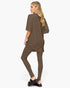 A woman modeling a matching two-piece lounge set consisting of an oversized short-sleeve tunic top and high-waisted leggings in a solid taupe-brown color, shown from a three-quarters back view against a light gray background.