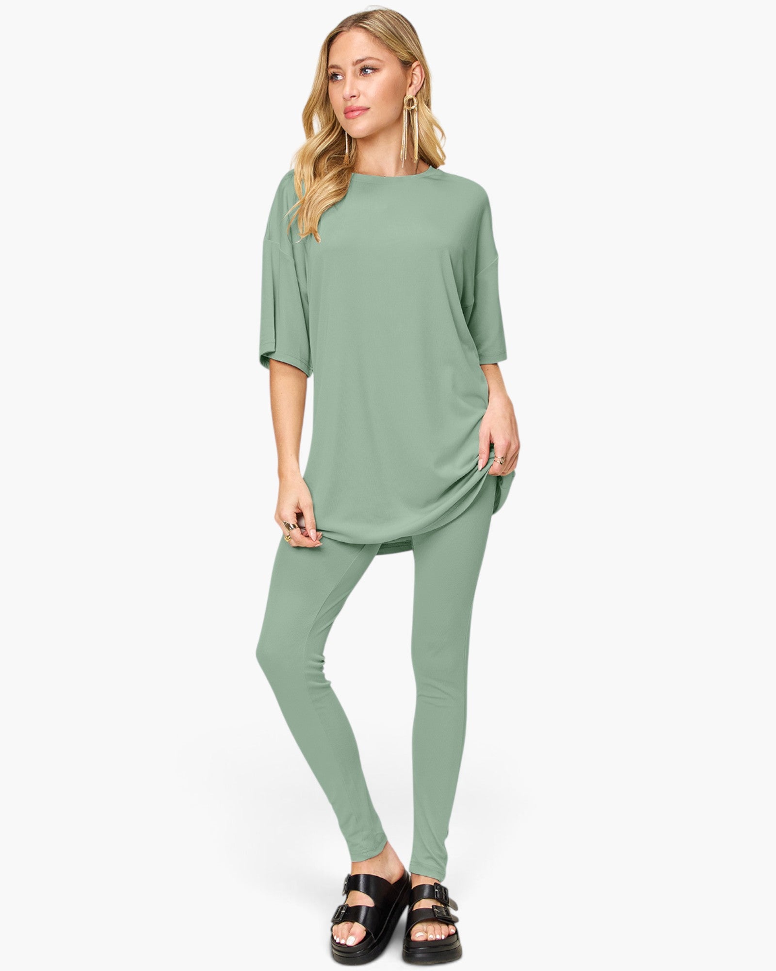 A woman modeling a sage green two-piece lounge set consisting of a short-sleeve tunic top and matching leggings, paired with black slide sandals, against a plain white studio background.
