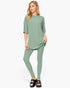 A woman modeling a sage green two-piece lounge set consisting of a short-sleeve tunic top and matching leggings, paired with black slide sandals, against a plain white studio background.