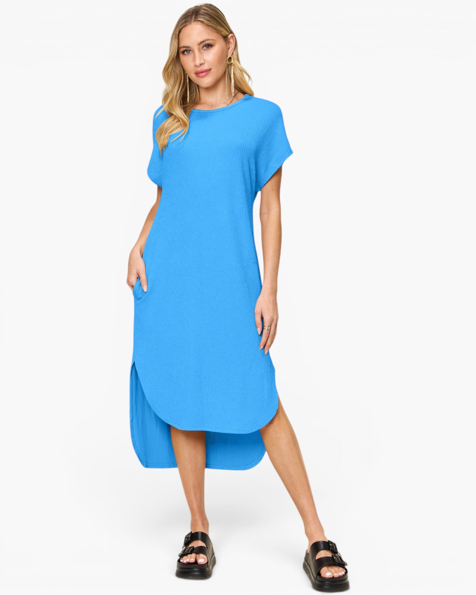 A woman modeling a bright blue short-sleeve midi dress with a high-low hem and side pockets, paired with black slide sandals, against a plain white studio background.