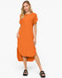 A woman modeling a loose-fitting orange short-sleeve midi dress with an asymmetrical hem, paired with black slide sandals, against a plain white studio background.