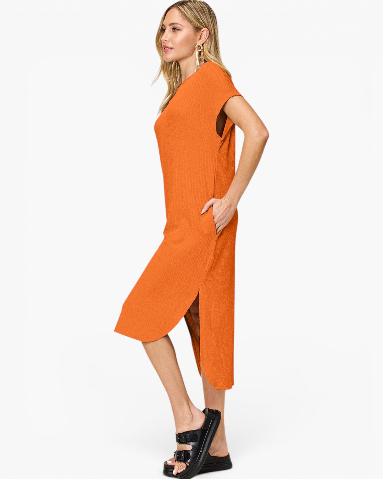 A woman modeling a bright orange midi dress with a high-low hem and side slit, paired with black platform sandals, against a plain white studio background.
