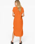 A woman shown from the back wearing a loose, short-sleeve orange midi dress with a side slit, paired with black loafers, against a plain white studio background.