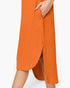 Close-up of a person wearing a bright orange ribbed midi dress with a side slit and hand tucked into the side pocket, against a plain light background.
