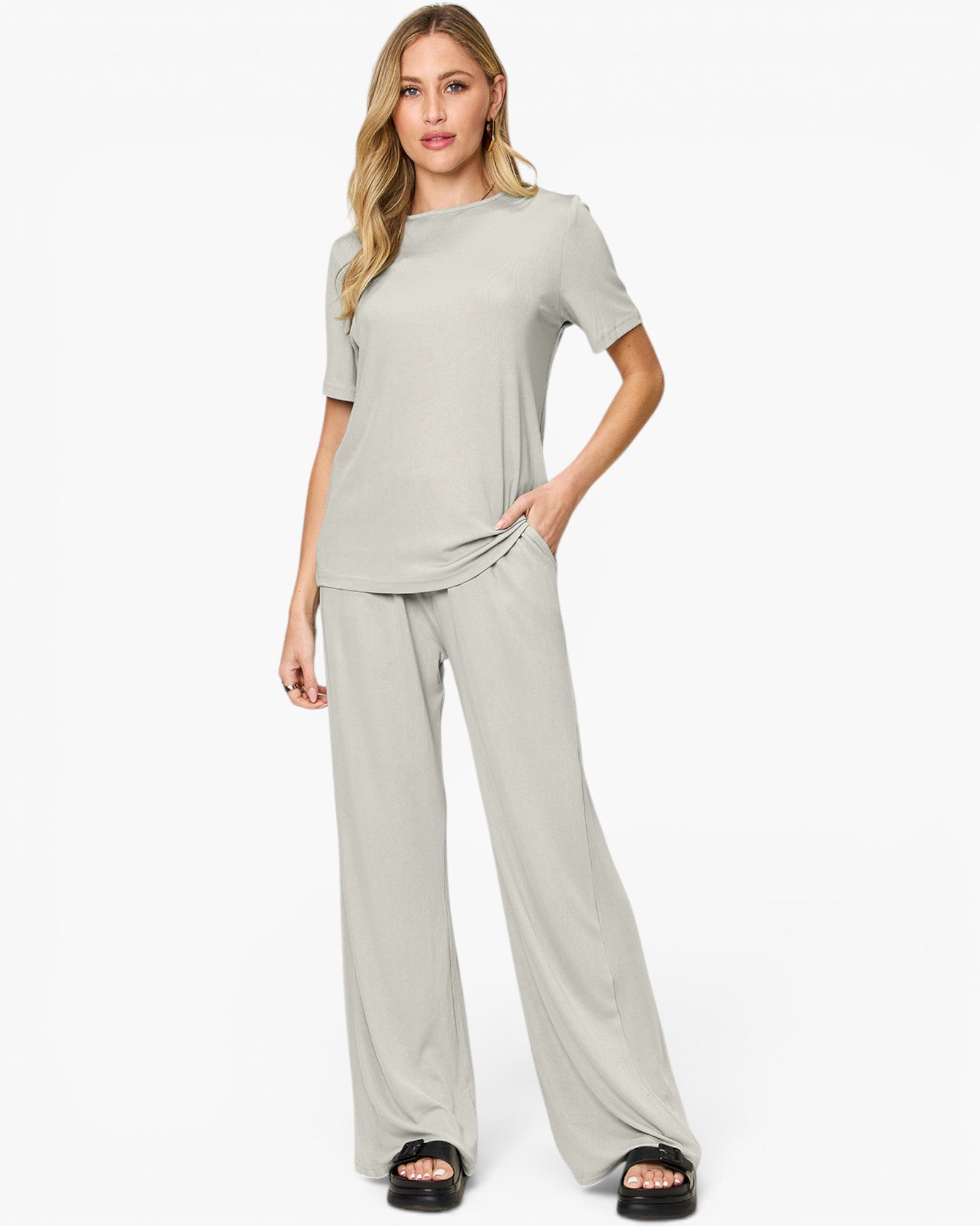 A woman modeling a light sage green two-piece lounge set consisting of a short-sleeve crewneck top and matching wide-leg pants, paired with black slide sandals against a plain white studio background.