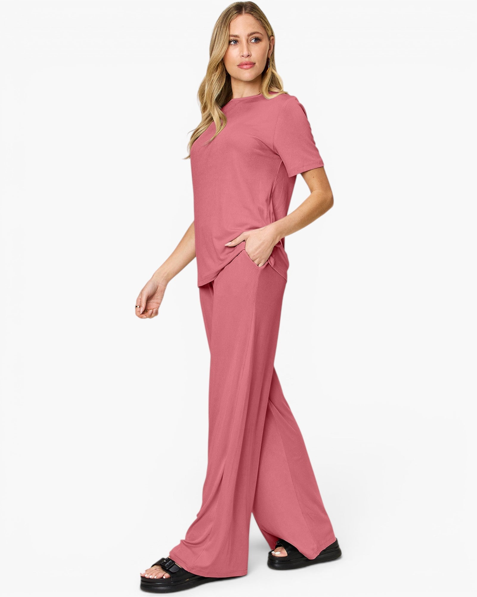 A woman modeling a dusty rose two-piece lounge set consisting of a short-sleeve top and matching wide-leg pants, paired with black slide sandals against a plain white studio background.