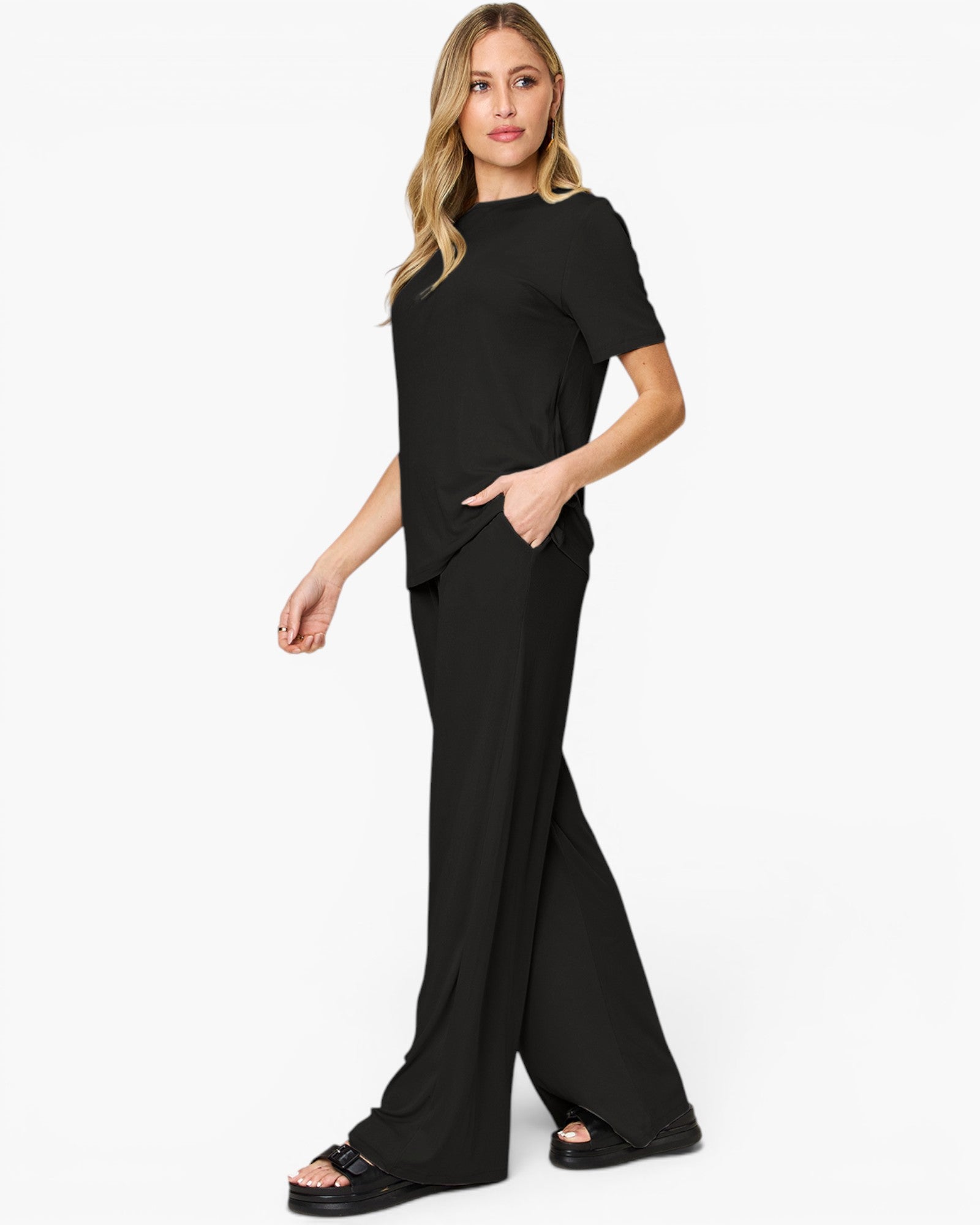 A woman modeling a black short-sleeve jumpsuit with wide-leg pants and side pockets, paired with black platform sandals, against a plain white studio background.