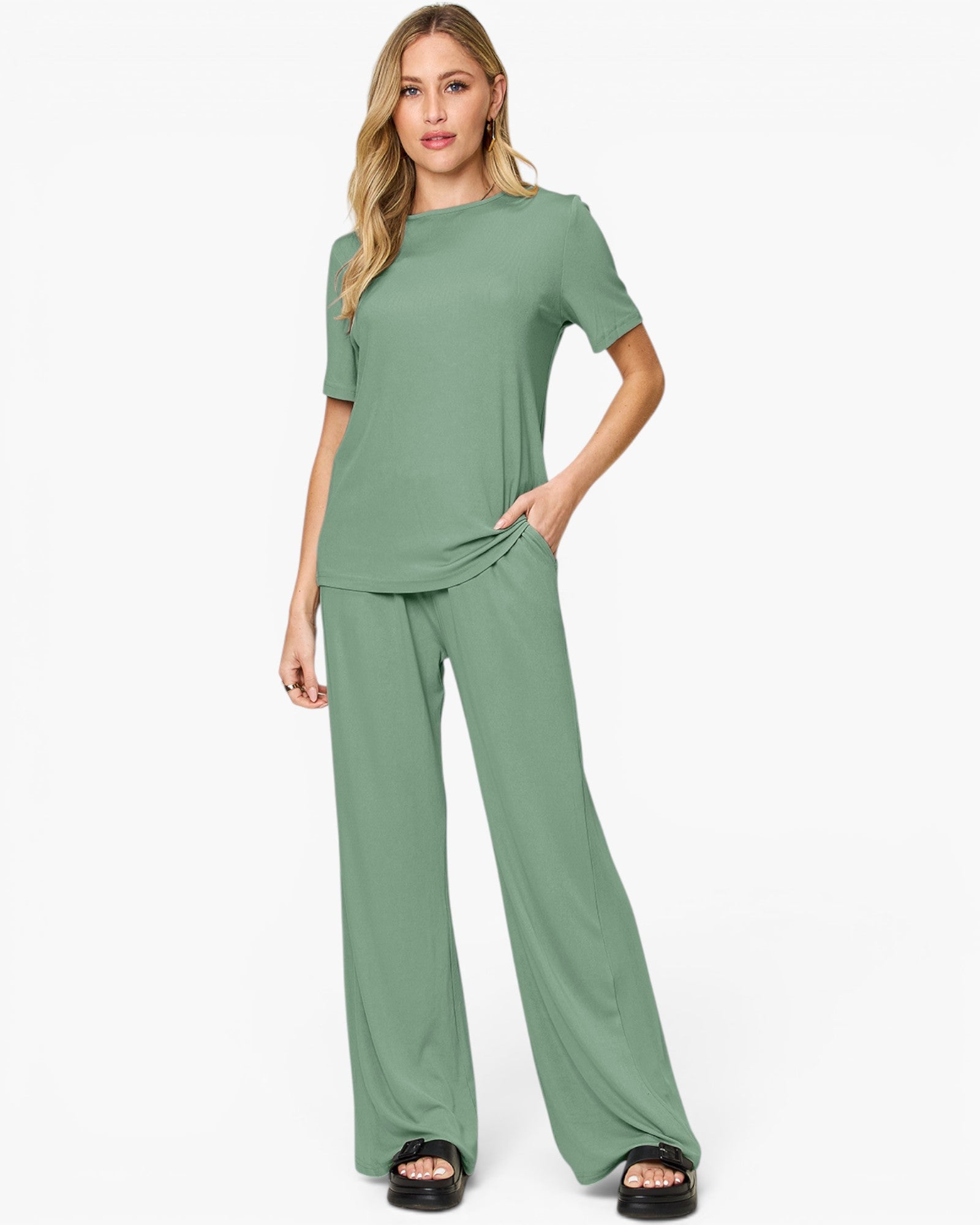 A woman modeling a sage green two-piece lounge set consisting of a short-sleeve crewneck top and matching wide-leg pants, paired with black slide sandals against a plain white studio background.