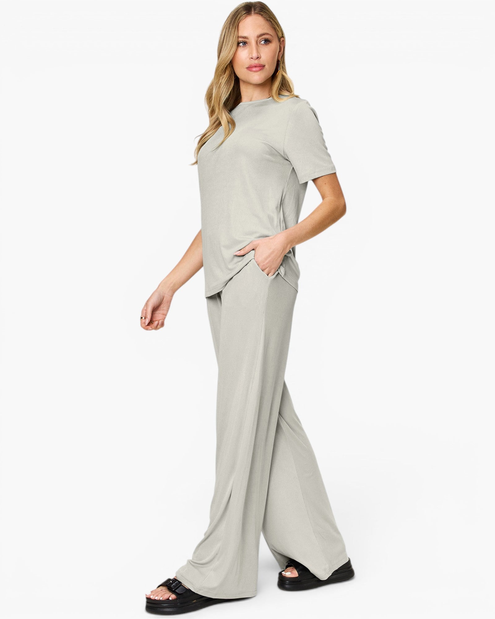 A woman modeling a light sage green two-piece lounge set consisting of a short-sleeve crewneck top and matching wide-leg pants, paired with black slide sandals against a plain white studio background.
