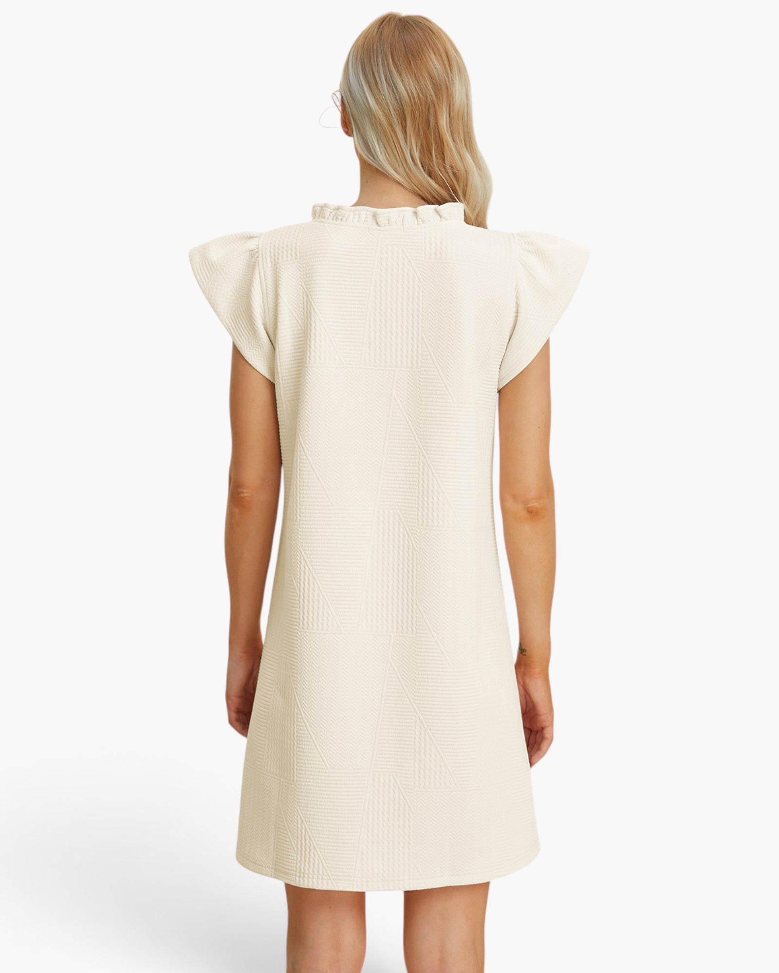 A woman viewed from the back wearing a cream-colored textured dress with ruffled shoulder details, set against a plain light gray studio background.