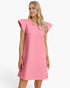 A woman modeling a pink textured shift dress with ruffled trim at the V-neck and cap sleeves, photographed against a clean white studio background.