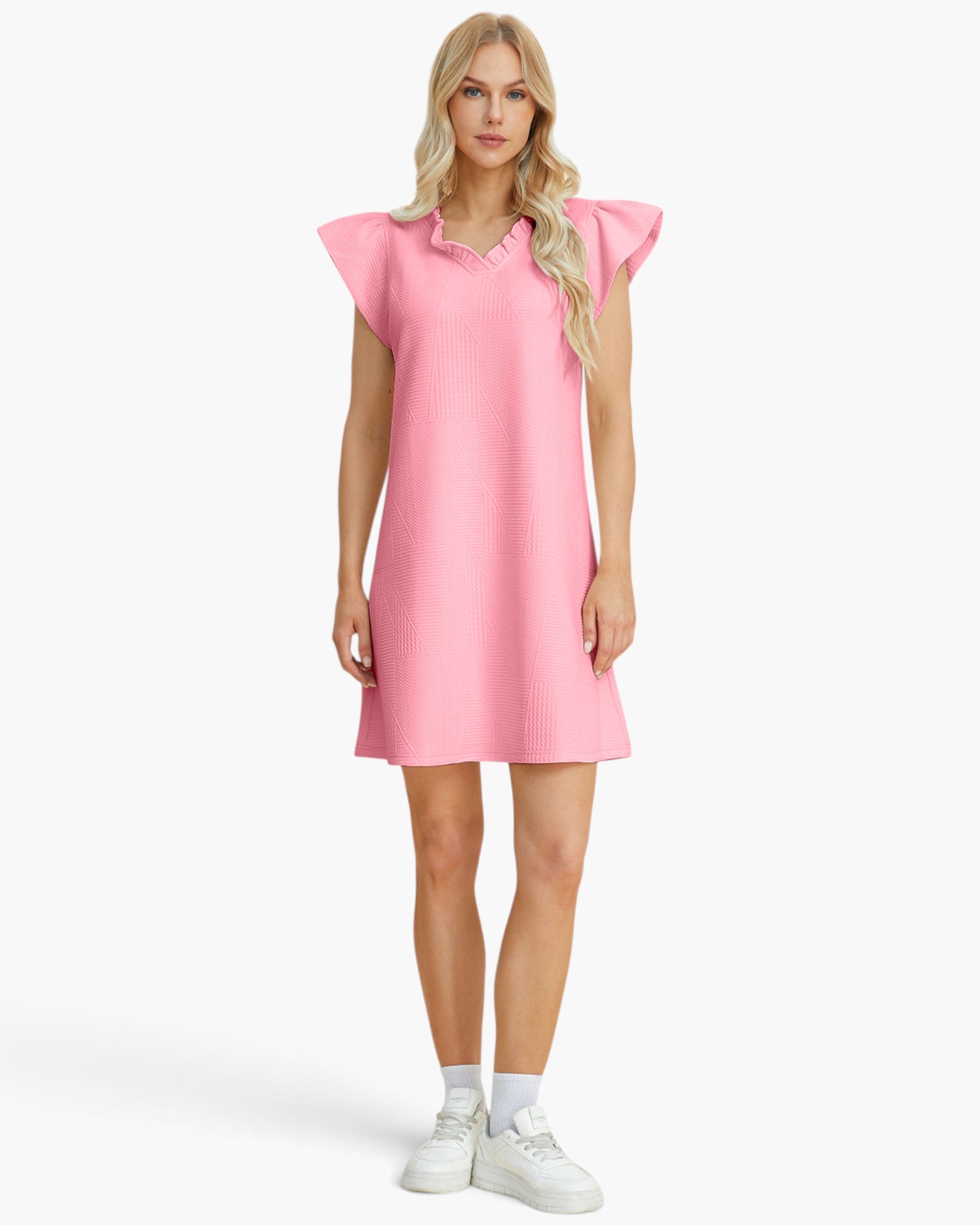 A woman modeling a light pink ribbed mini dress with ruffled cap sleeves and a ruffled collar, styled with white socks and white sneakers against a light gray studio background.