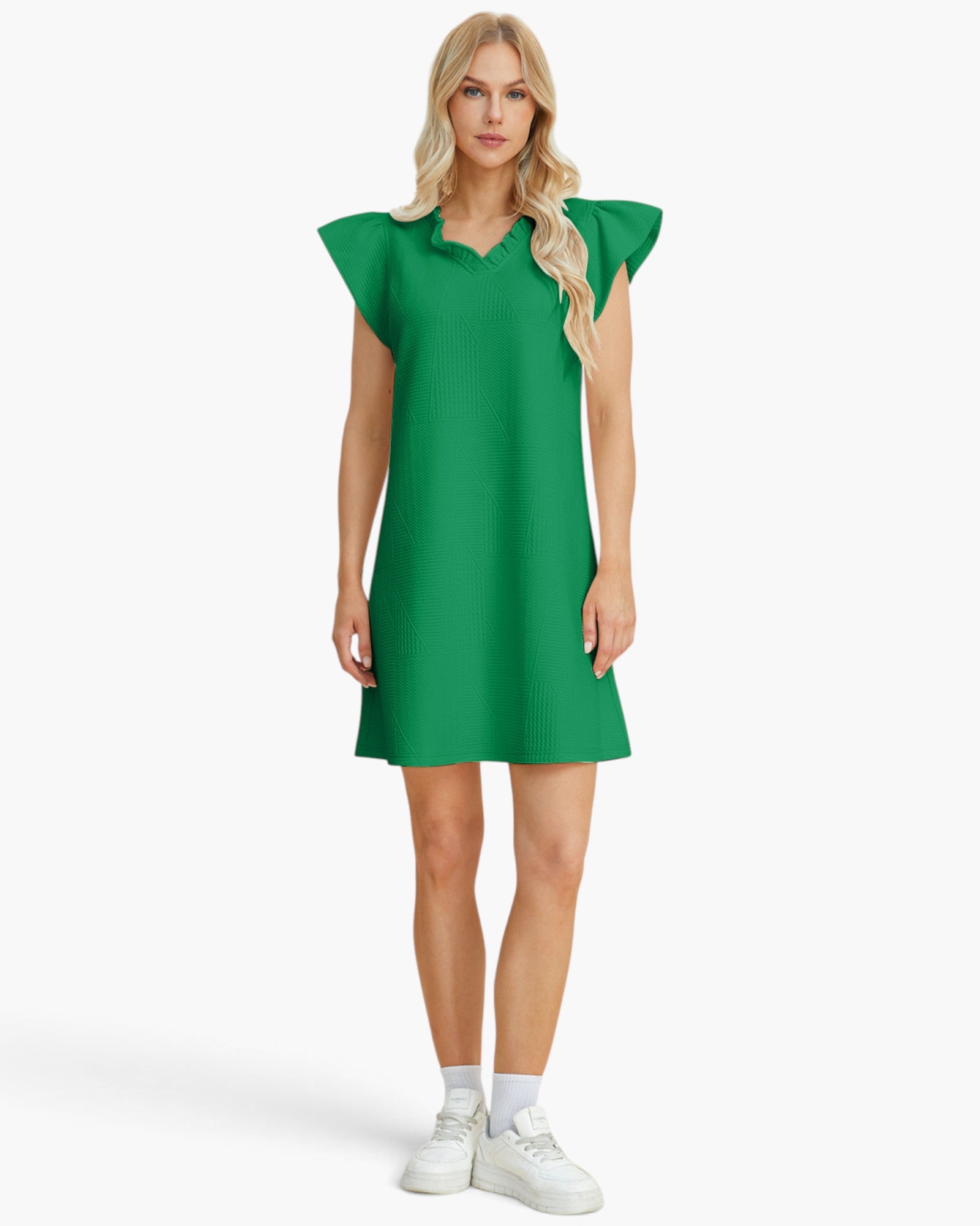 A blonde model wearing a bright green short-sleeve mini dress with ruffled cap sleeves, white socks, and white sneakers, standing against a light gray studio background.