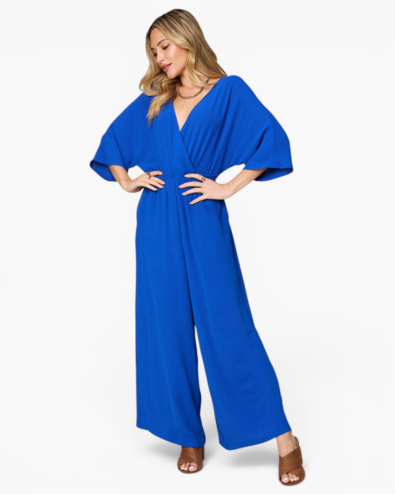 A woman modeling a bright royal blue wide-leg jumpsuit with a wrap-style V-neck bodice and voluminous elbow-length sleeves, photographed against a clean light gray studio background.