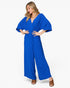 A woman modeling a bright royal blue wide-leg jumpsuit with a wrap-style V-neck bodice and voluminous elbow-length sleeves, photographed against a clean light gray studio background.