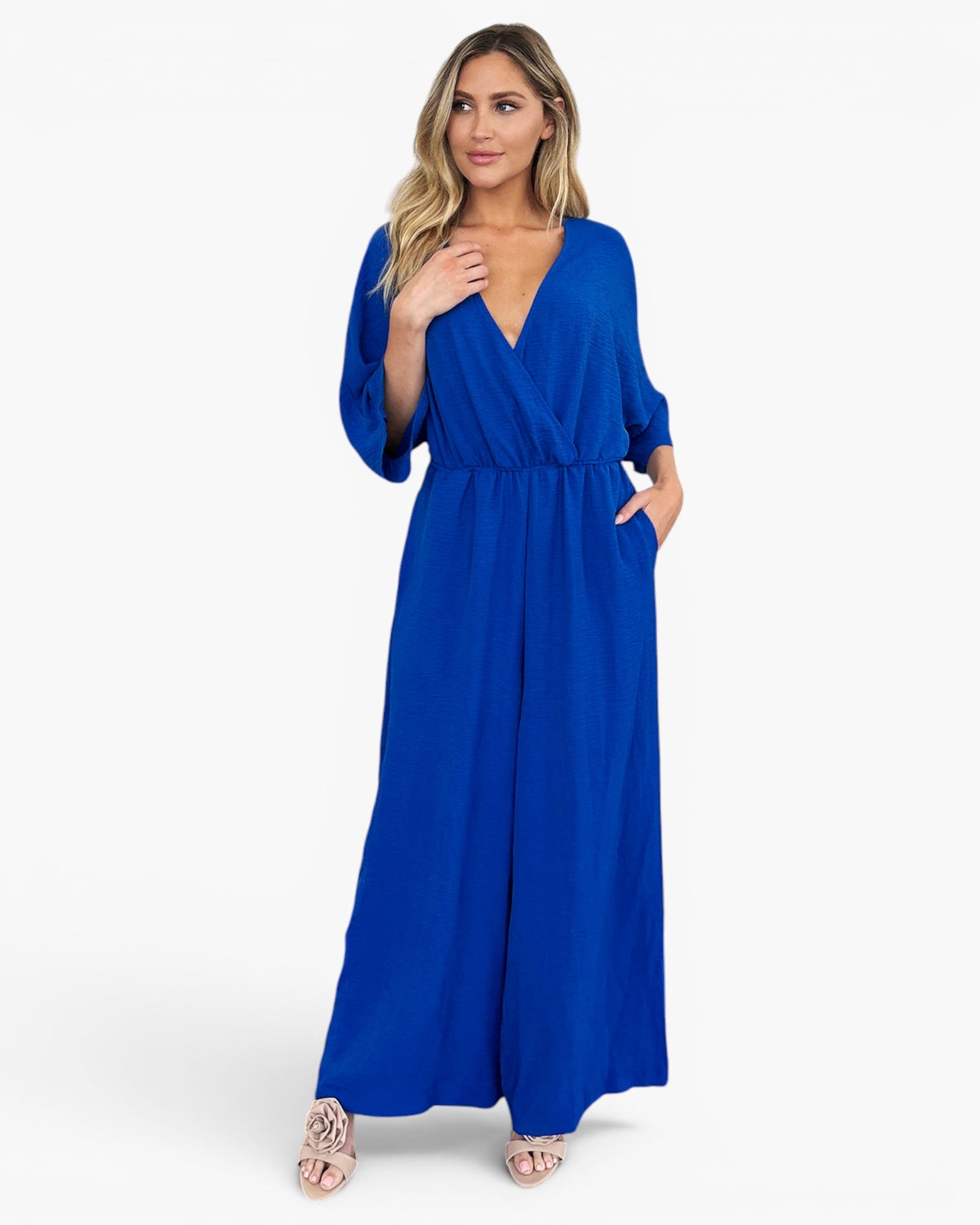 A woman modeling a royal blue maxi jumpsuit with a wrap-style V-neck, elbow-length sleeves, side pockets, and a tie waist, photographed against a clean white studio background.