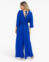 A woman modeling a royal blue wide-leg jumpsuit from the back, featuring a deep V-neckline with tie-back straps, three-quarter sleeves, and a drawstring waist, set against a plain white studio background.