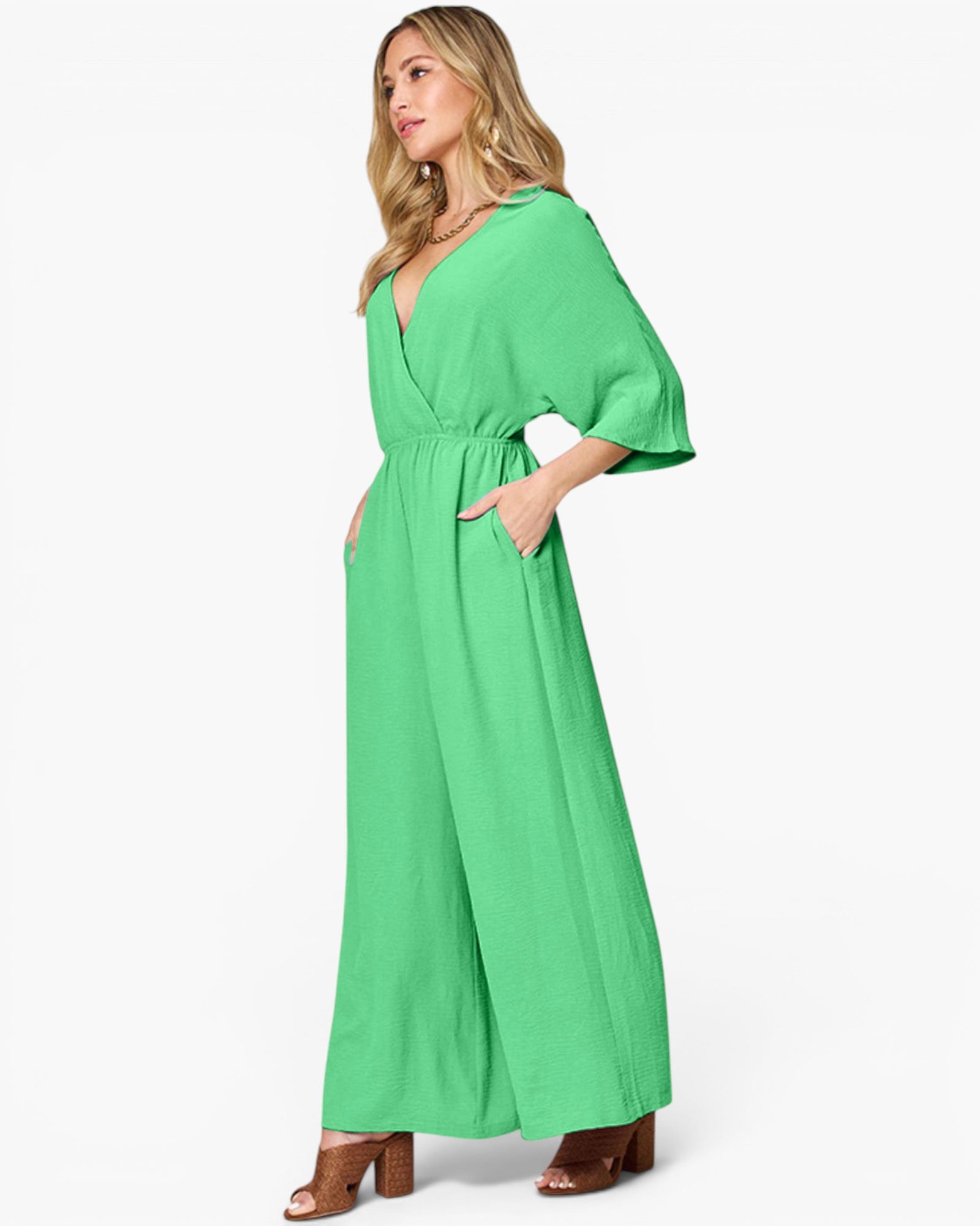 A woman modeling a bright green wide-leg jumpsuit with three-quarter sleeves and a wrap-style V-neckline, shown against a plain light gray studio background.