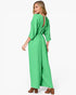 A woman modeling a bright green jumpsuit from the back, showing a draped open back and wide legs, paired with brown block-heel sandals against a plain white background.