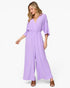 A woman modeling a lavender-purple wide-leg jumpsuit with a wrap-style V-neck bodice and elbow-length sleeves, paired with brown heeled sandals against a plain white studio background.